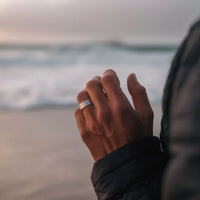 Primer plano de una mano que lleva el Anillo de Magnoterapia - Alivio para la menopausia, con las olas del mar de fondo. La persona, vestida con una chaqueta oscura, disfruta de la playa al atardecer o al amanecer.